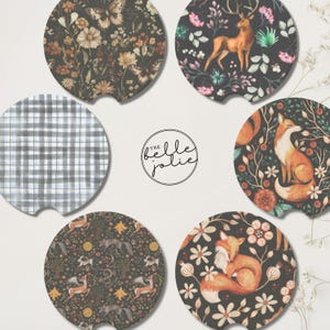 May include: Six circular car cup holder coasters featuring diverse designs: one with butterflies and flowers, one with a deer in a forest, one with a grey plaid pattern, one with a fox, one with woodland creatures, and one with a sleeping fox. Text reads the belle jolie.