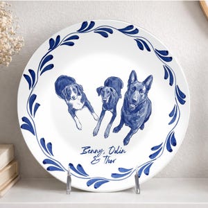 May include: A white ceramic plate featuring a blue painted border of stylized leaves. The center displays three dog illustrations in blue ink, with the text "Benny, Odin & Thor" printed below the dogs in a cursive blue script font.