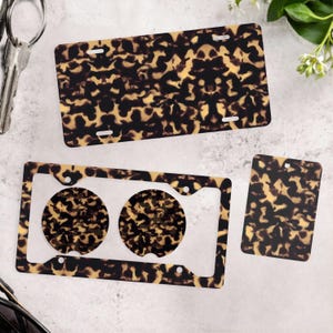 May include: A rectangular license plate frame, a license plate cover with four mounting slots, and a rectangular air freshener, all featuring a brown and black tortoiseshell pattern. The license plate cover includes two circular cutouts.