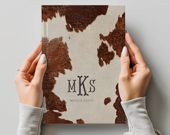 May include: A rectangular notebook featuring a brown and white cowhide print pattern. The center of the cover displays the dark brown monogram letters MKS in a stylized font, with the name Maggie Kelly printed in smaller text below.