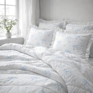 May include: A white quilted bedding set featuring a repeating pattern of light blue bows. The set includes a comforter and matching pillow shams, each finished with a ruffled edge. The fabric is white with a soft, light blue bow print throughout.