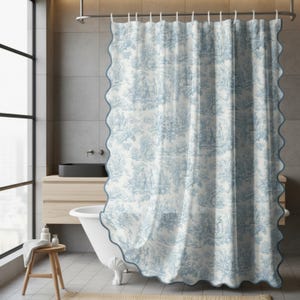 May include: A blue and white toile shower curtain with a scalloped edge. The curtain features a repeating pattern of pastoral scenes. The curtain is hanging in a modern bathroom with a white bathtub and wooden stool.