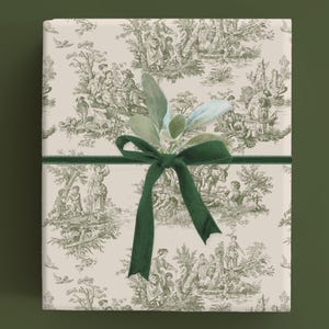 May include: A cream-colored gift wrap with a green and white toile pattern featuring a pastoral scene. The wrap is tied with a green velvet ribbon and a sprig of green foliage.