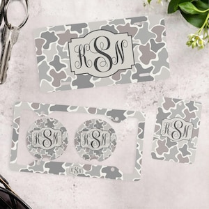 May include: A rectangular license plate, a license plate frame with two circular cup holder inserts, and a rectangular tag, all featuring a gray, white, and taupe cow print pattern with a central gray plaque displaying the monogram KSN.