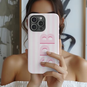 Preppy monogrammed phone case with retro vertical stripes in light pastel pink and white, custom initials ‘AKL’ in a bold shadow design for a trendy look. This personalized iPhone cover is made to protect your phone while adding a beachy, summer-inspired aesthetic. Great custom gift idea for birthdays, back to school, or sorority sisters. Available for latest iPhone models with more color options. Add your own initials for a unique touch that stands out from ordinary phone cases.