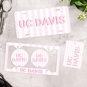 May include: A set of UC Davis accessories, including a license plate, car coasters, and a car air freshener. Each item features a pink and white striped pattern with the text "UC DAVIS" in a matching pink color.