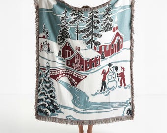 May include: A woven throw blanket featuring a winter landscape with red houses, a church, a stone bridge, and a winding blue river. The scene includes tall black pine trees and figures building a snowman on a white background with fringed edges.