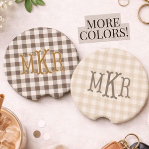 May include: Two car coasters with a gingham pattern and personalized initials. One coaster is brown and white, the other is beige and white. The text "MORE COLORS!" is displayed above the coasters.