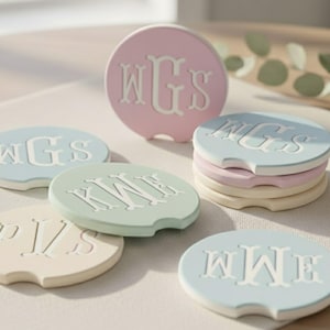 May include: A collection of pastel-colored car coasters with custom monogram designs. The coasters are round with a small notch and feature white, raised lettering. Colors include light pink, blue, green, and cream. The coasters are stacked and scattered.