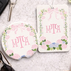 May include: A round ceramic car coaster and a rectangular hanging air freshener, both featuring a white background with a pink bow, green vines, and pink and blue hydrangea flowers. The monogram letters MTA are printed in pink in the center.