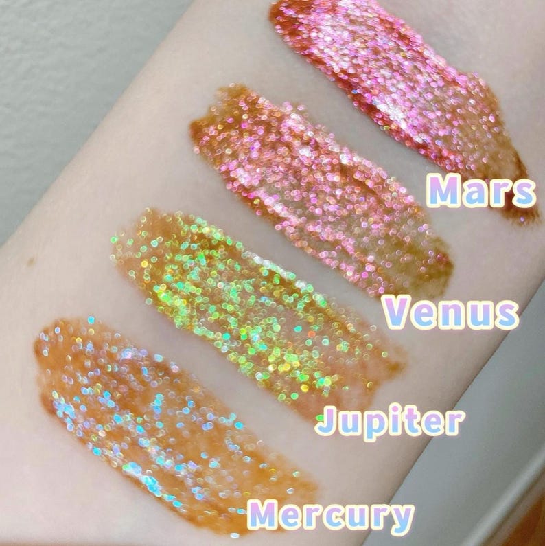 May include: Four swatches of iridescent glitter makeup on a person's arm. The swatches are labeled "Mercury", "Jupiter", "Venus", and "Mars".