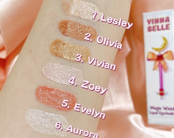 May include: Six different shades of liquid eyeshadow swatches on a person's arm. The shades are labelled 1. Lesley, 2. Olivia, 3. Vivian, 4. Zoey, 5. Evelyn, and 6. Aurora. The swatches are on a pink satin background. The image also includes a small bottle of liquid eyeshadow with the brand name "Vinna Belle" and the words "Magic Wand Liquid Eyeshadow".