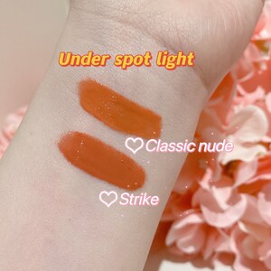 May include: Two swatches of lipstick on a person's arm. The top swatch is labeled "Classic nude" and the bottom swatch is labeled "Strike". The text "Under spot light" is written above the swatches.