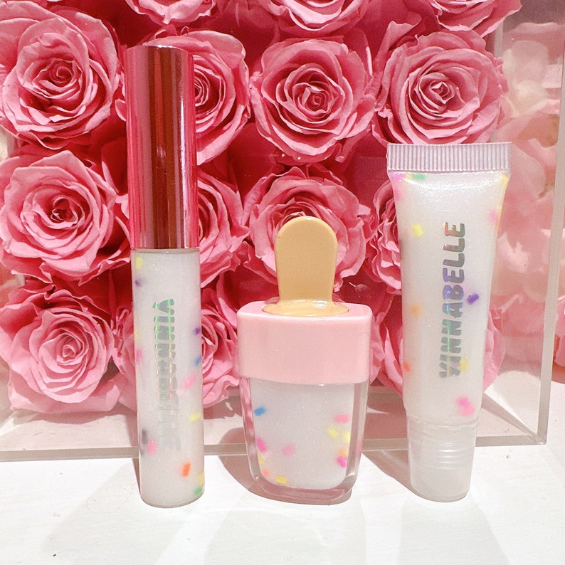 May include: Three clear lip glosses with colorful sprinkles. The glosses are in different containers: a tube, a popsicle-shaped container, and a squeeze tube. The squeeze tube has the text "VINNABELLA" on it.