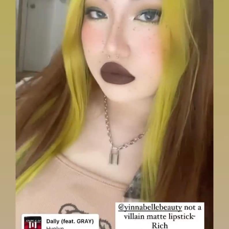 May include: A person with yellow and brown hair, wearing a silver chain necklace with a padlock pendant. The person is wearing dark brown lipstick and has a nose ring. The text on the image says "@yinnabellebeauty not a villain matte lipstick - Rich".