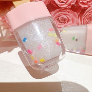 May include: A clear plastic tube filled with a white, glittery lip gloss. The gloss has colorful sprinkles throughout. The tube is topped with a pink lid.