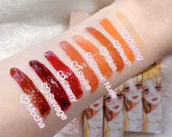 Pode incluir: Um close-up de dez tons diferentes de gloss labial aplicados no braço de uma pessoa. Os tons variam de um marrom escuro a um pêssego claro. Os gloss labiais são etiquetados com nomes como "Mocha", "Savage", "Cherry", "Strike", "Classic Nude", "Skin", "Peach" e "Darling".