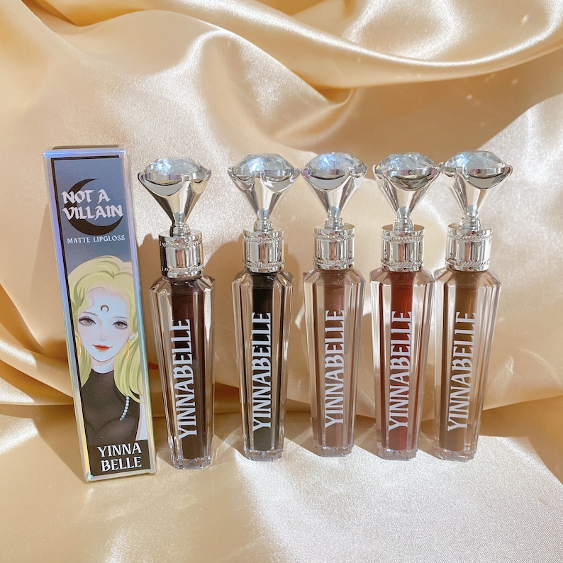 May include: Matte lipgloss collection by YINNABELLE. The bottles have diamond-shaped tops and are arranged with a box that reads "NOT A VILLAIN" and features a cartoon illustration. The lipgloss shades vary from dark brown to reddish-brown.