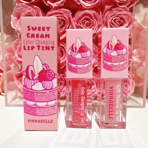 K&ouml;nnte beinhalten: Drei rosa Lipgloss-Tuben mit einer Farbe wechselnder Formel. Die Tuben zeigen eine Cartoon-Illustration eines rosa und wei&szlig;en mehrschichtigen Kuchens mit Erdbeeren und Schlagsahne. Der Text auf den Tuben lautet "Color Changing Lip Tint" und "VINNABELLE".