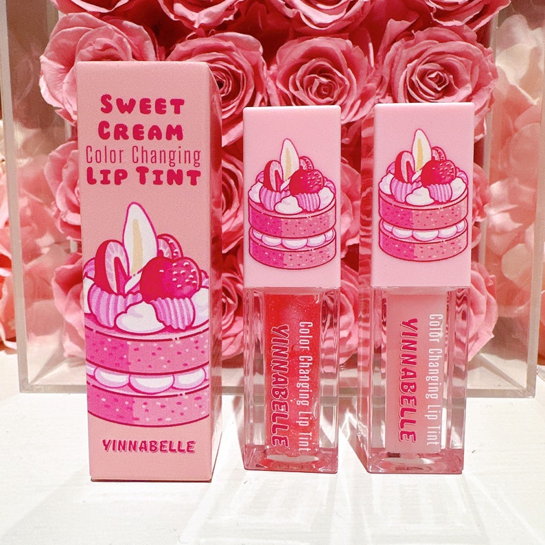 K&ouml;nnte beinhalten: Drei rosa Lipgloss-Tuben mit einer Farbe wechselnder Formel. Die Tuben zeigen eine Cartoon-Illustration eines rosa und wei&szlig;en mehrschichtigen Kuchens mit Erdbeeren und Schlagsahne. Der Text auf den Tuben lautet "Color Changing Lip Tint" und "VINNABELLE".
