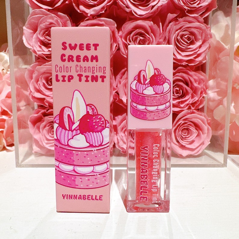 K&ouml;nnte beinhalten: Ein rosa und wei&szlig;er Lip Tint-Stift mit einer Farbe wechselnder Formel. Der Stift zeigt eine Comic-Illustration eines rosa und wei&szlig;en mehrst&ouml;ckigen Kuchens mit Erdbeeren und Schlagsahne. Der Text auf dem Stift lautet "Sweet Cream Color Changing Lip Tint" und "Yinnabelle".
