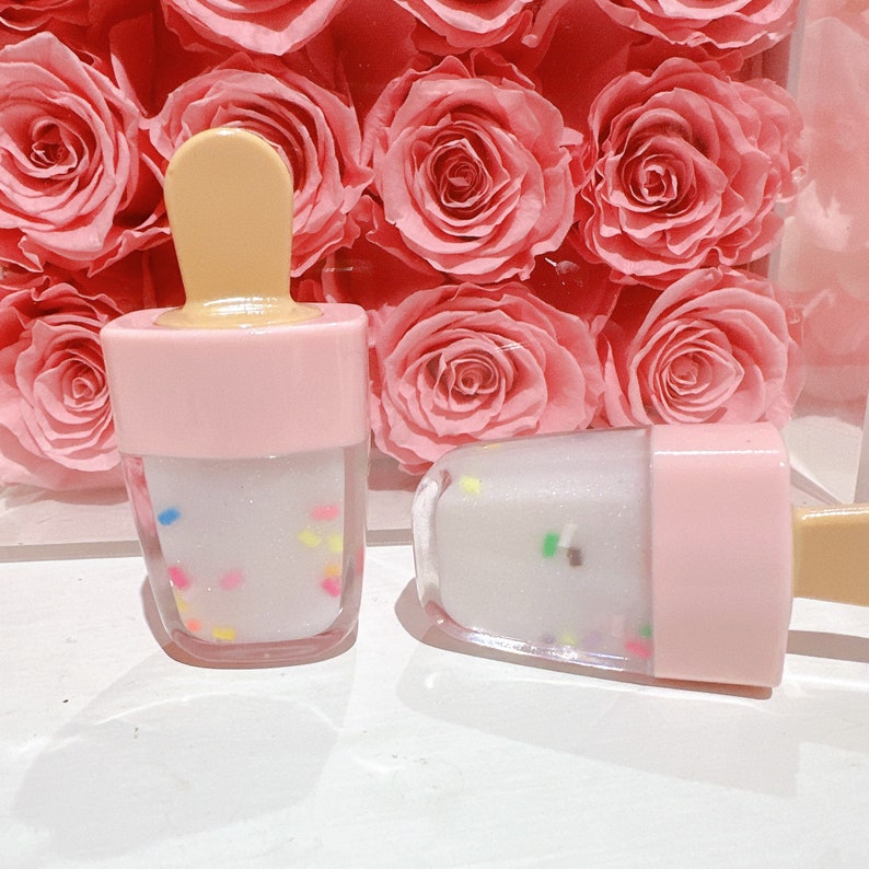 May include: Two pink and white popsicle-shaped lip gloss tubes with a clear, glittery gloss and colorful sprinkles inside.
