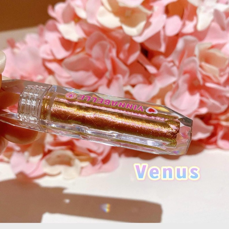 May include: A clear, crystal-shaped tube of gold glitter lip gloss with the text "VINNABELLE" in pink lettering. The tube is labeled "Venus".
