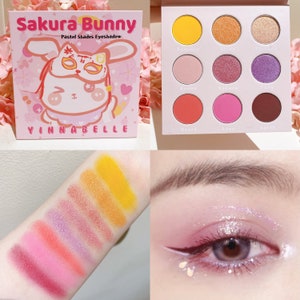 May include: A pastel eyeshadow palette with a cartoon bunny design. The palette has nine shades, including yellow, orange, pink, and purple. The palette is labeled "Sakura Bunny Pastel Shades Eyeshadow" and "Yinnabelle".