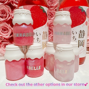 K&ouml;nnte beinhalten: F&uuml;nf kleine Glasflaschen mit rosa und wei&szlig;em Glitzer-Lippenbalsam. Die Flaschen sind in Form von Milchpackungen und haben wei&szlig;e Deckel mit rosa Schleifen. Die Flaschen sind mit dem Text "VINNABEL" und "ABELLE" beschriftet.