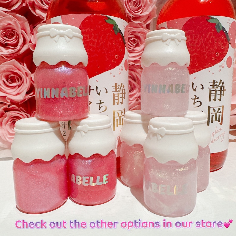 K&ouml;nnte beinhalten: F&uuml;nf kleine Glasflaschen mit rosa und wei&szlig;em Glitzer-Lippenbalsam. Die Flaschen sind in Form von Milchpackungen und haben wei&szlig;e Deckel mit rosa Schleifen. Die Flaschen sind mit dem Text "VINNABEL" und "ABELLE" beschriftet.