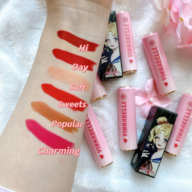 May include: Six pink lipstick tubes with gold accents and a Sailor Moon character on the packaging. The lipsticks are labeled "Hi", "Day", "Soft", "Sweets", "Popular", and "Charming".