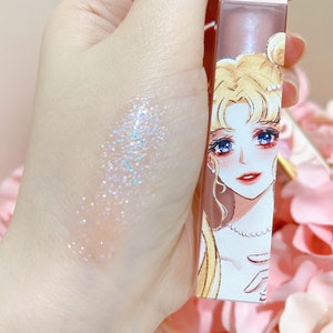May include: A clear lip gloss with iridescent glitter in a tube featuring a cartoon illustration of a woman with blonde hair and a pearl necklace. The tube is pink and white with the text "Sailor Moon" on the side.