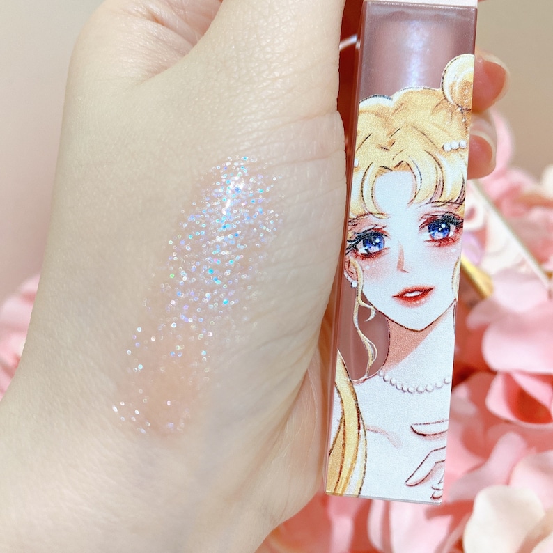 May include: A clear lip gloss with iridescent glitter in a tube featuring a cartoon illustration of a woman with blonde hair and a pearl necklace. The tube is pink and white with the text "Sailor Moon" on the side.
