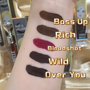 May include: A close-up of a person's arm displaying five lipstick swatches in shades of brown, burgundy, and black. The names "Boss Up", "Rich", "Bloodshot", "Wild", and "Over You" are written next to each swatch. The image also shows lipstick tubes.