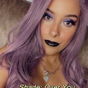 May include: A woman with lavender hair and makeup, including dark lipstick and eye shadow. She wears a silver necklace with a heart pendant. The text "Shade: Over You" is displayed in a yellow font.