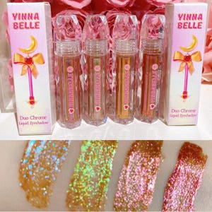 May include: Three clear bottles of liquid eyeshadow with a pink and gold glitter finish. The bottles are labeled "Vinna Belle" and "Duo Chrome Liquid Eyeshadow". The bottles are on a white surface with pink flowers in the background.