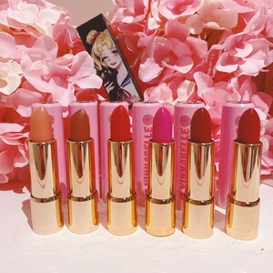 May include: Six pink and gold lipstick tubes with the word "VINARELLA" printed on the side. The lipsticks are arranged in a row on a white surface.