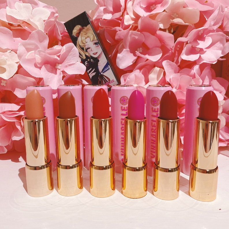 May include: Six pink and gold lipstick tubes with the word "VINARELLA" printed on the side. The lipsticks are arranged in a row on a white surface.