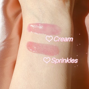 K&ouml;nnte beinhalten: Zwei Lippgloss-Swatches auf dem Arm einer Person. Der obere Swatch ist mit "Cream" beschriftet und der untere Swatch mit "Sprinkles".