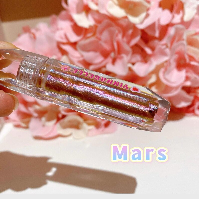 May include: A clear, iridescent lip gloss tube with a pink and white label that reads "VINNABELLA" and a heart symbol. The lip gloss is a shimmery, bronze color. The text "Mars" is written in a pink and blue gradient below the tube.
