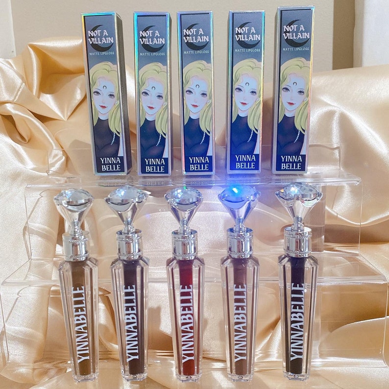 May include: Five YINNABELLE matte lipgloss tubes with diamond-shaped tops are displayed. The lipgloss tubes are arranged on a clear acrylic shelf. Each tube has a different shade of brown and red. The boxes above the tubes read "NOT A VILLAIN".