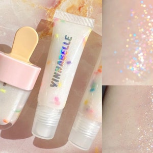 May include: Two clear lip gloss tubes with a white background and colorful sprinkles. The lip gloss tubes are labeled "Vinnarelle". One tube is in a popsicle shape.