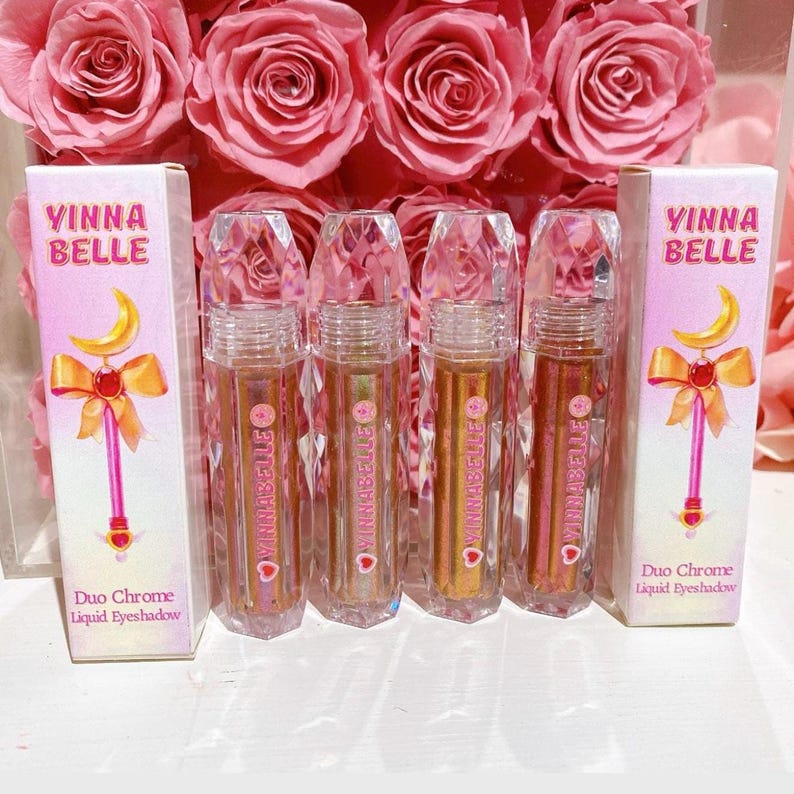May include: Four clear bottles of liquid eyeshadow with a pink and gold glitter effect. The bottles are labeled "Vinna Belle" and "Duo Chrome Liquid Eyeshadow".