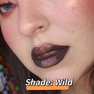May include: Close-up of a person's face with dark, metallic lipstick. The lipstick has a deep brown base with bronze shimmer. The text "Shade: Wild" is displayed in a white font on an orange background.