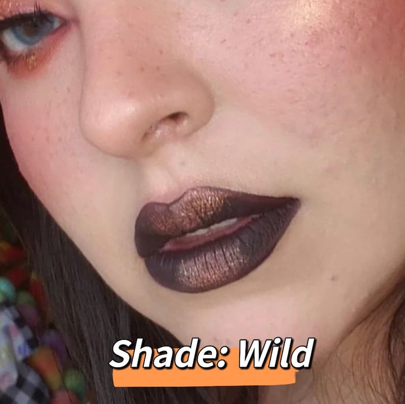 May include: Close-up of a person's face with dark, metallic lipstick. The lipstick has a deep brown base with bronze shimmer. The text "Shade: Wild" is displayed in a white font on an orange background.