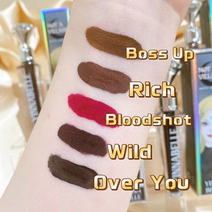 May include: A hand displays five swatches of matte liquid lipstick in shades of brown, red, and dark purple. The names of the shades are Boss Up, Rich, Bloodshot, Wild, and Over You. The lipstick tubes have a diamond-shaped cap.