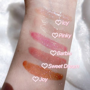 May include: Five different shades of lip gloss with glitter, swatched on a person's arm. The shades are labeled "Icy", "Pinky", "Barbie", "Sweet Dream", and "Joy".