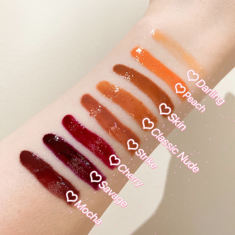 May include: A close-up of a hand with swatches of eight different shades of lip gloss. The shades are labeled from lightest to darkest: Darling, Peach, Skin, Classic Nude, Strike, Cherry, Savage, and Mocha.
