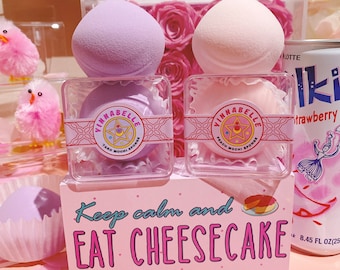 Puede incluir: Dos esponjas de maquillaje rosas y moradas en recipientes transparentes con el texto "VINNABELLE" y "Taro Mochi Sponge" y "Peach Mochi Sponge". Los recipientes están sobre una superficie rosa con un cartel que dice "Keep calm and EAT CHEESECAKE".