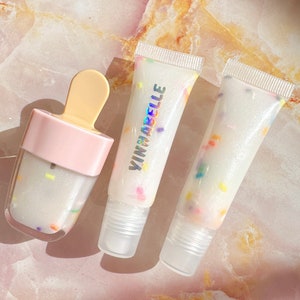 May include: Three tubes of clear lip gloss with colorful sprinkles inside. The lip gloss is in a clear tube with a white cap. The middle tube has the brand name "VINMACELLE" printed on the side.